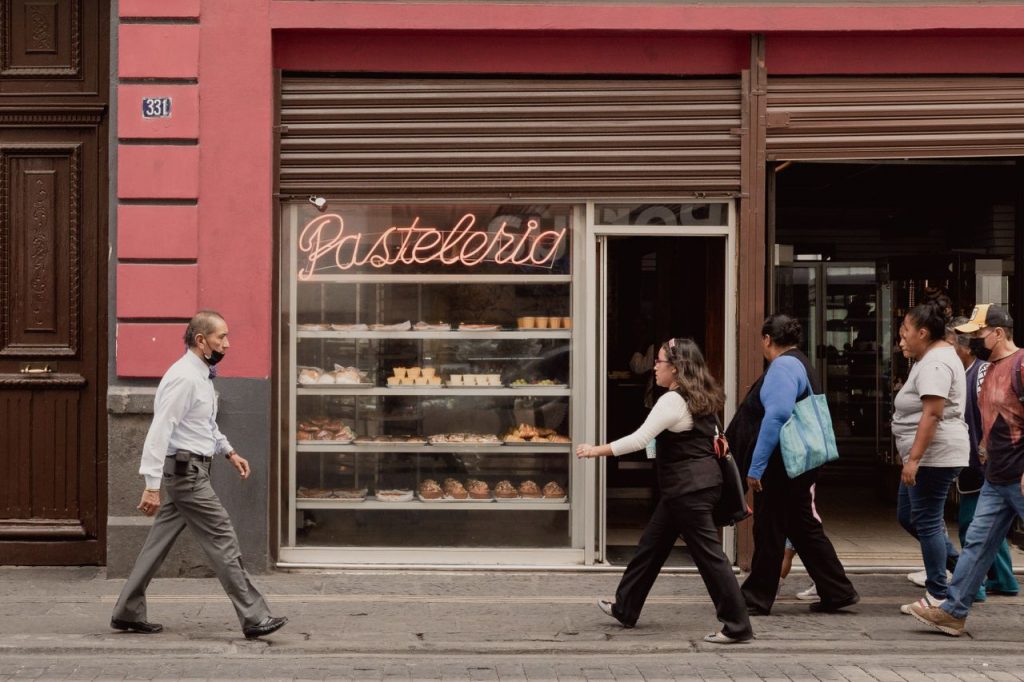 Pedestrians passing by a bakery storefront with neon signage in Puebla, Mexico.