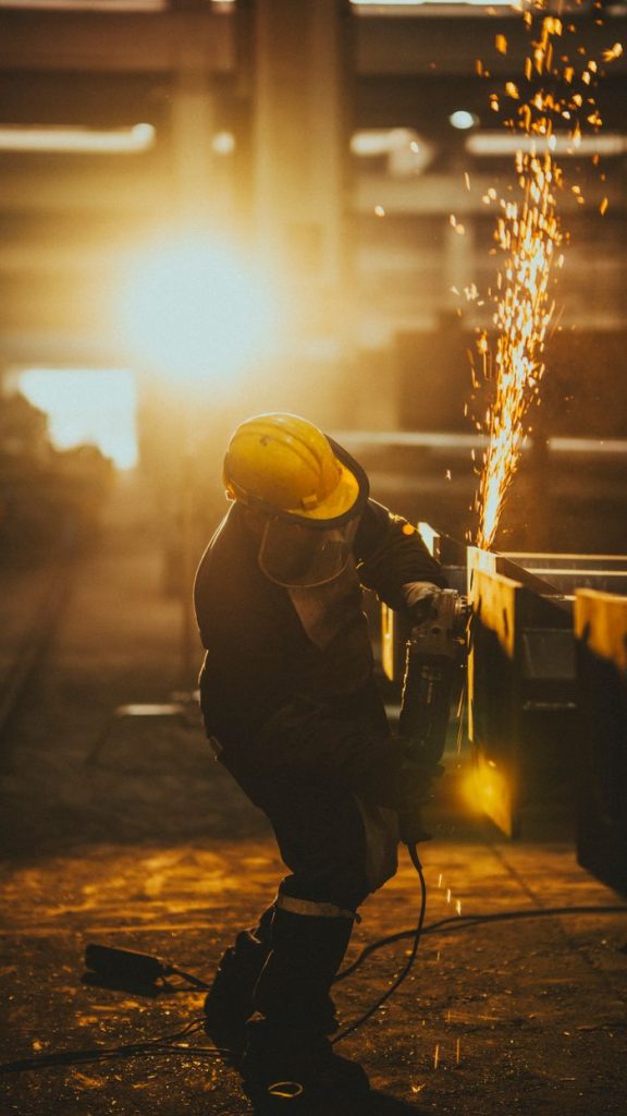 Silhouette of a worker in a hard hat, using a grinder to cut metal, with sparks flying amidst golden sunlight.